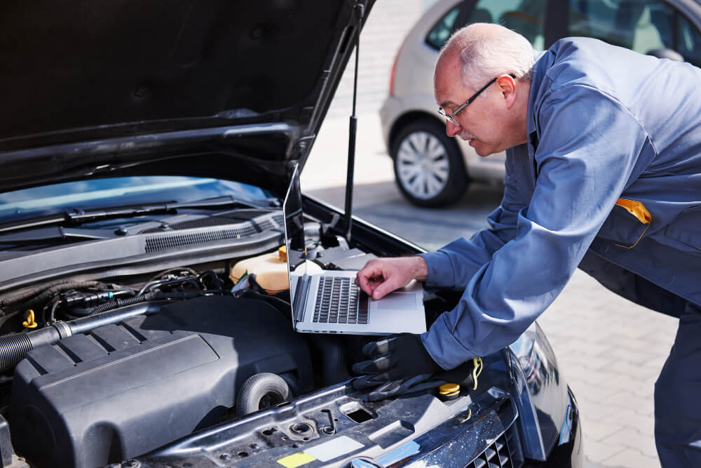 Ein Mechaniker in Arbeitskleidung prüft mithilfe eines Laptops ein Unfallgutachten und inspiziert dabei den geöffneten Motorraum eines Autos
