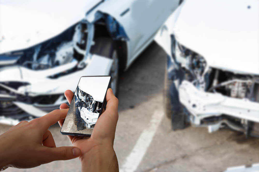 Eine Person fotografiert mit dem Smartphone zwei beschädigte weiße Autos nach einem Unfall zur Erstellung eines Kfz-Gutachtens