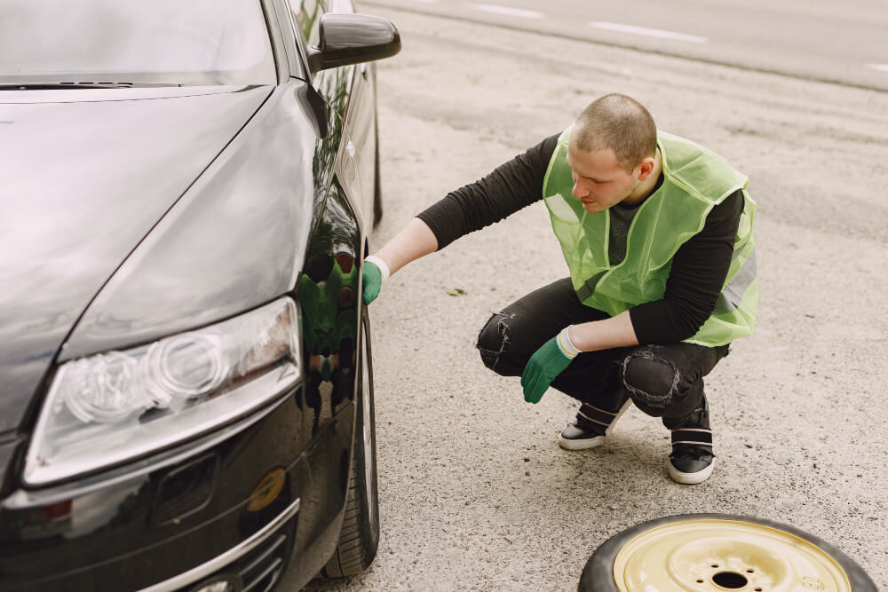 Ein Mann mit grüner Warnweste und Handschuhen prüft einen platten Reifen, als wäre es eine automatisch gespeicherte Panne