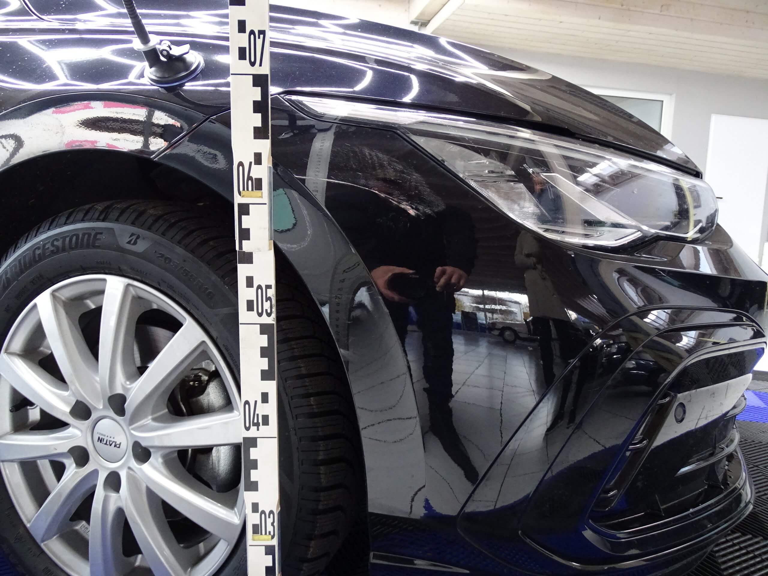 Nahaufnahme der Vorderseite eines schwarzen Autos mit Messstab neben dem Rad, in dem sich die fotografierende Person spiegelt