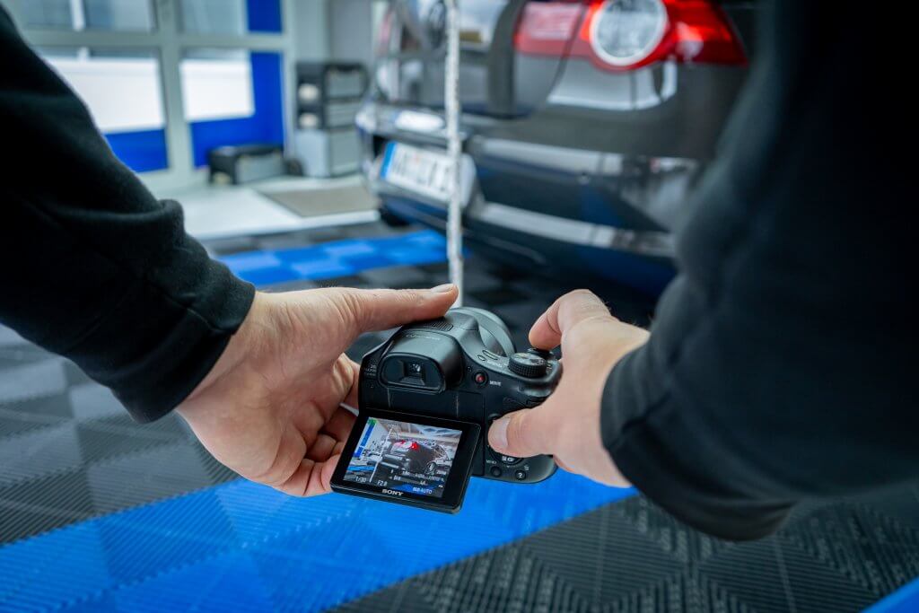 Eine Person fotografiert mit einer Kamera das Heck eines schwarzen Autos in einer Garage mit blau-schwarzem Boden