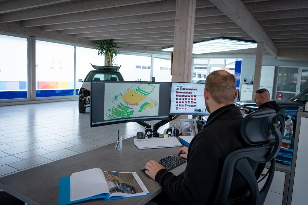 Ein Mann arbeitet an einem Schreibtisch im modernen Büro, betrachtet ein 3D-Automodell am Bildschirm, draußen steht ein Auto vor großen Fenstern