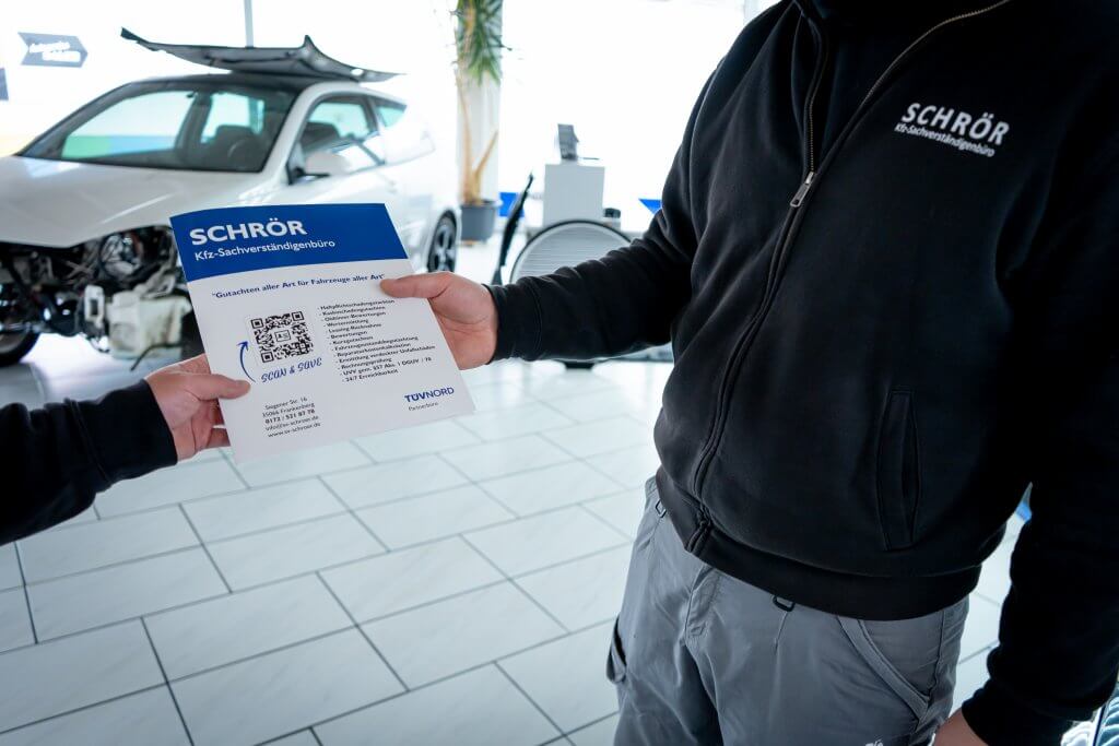 In einer Autowerkstatt tauschen zwei Personen einen Flyer aus, eine trägt eine Jacke mit dem Schriftzug SCHRÖR