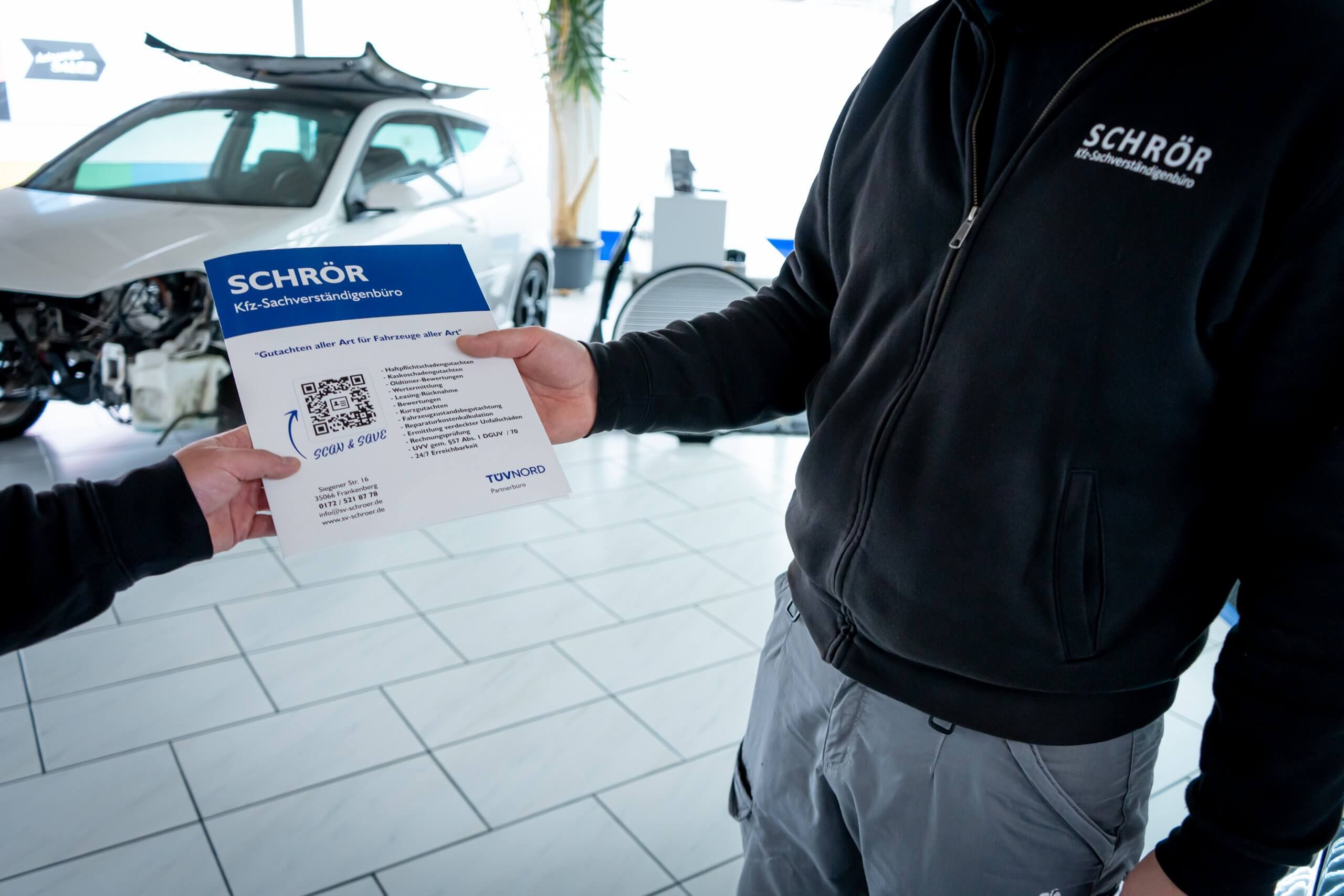 In einer Autowerkstatt tauschen zwei Personen einen Flyer aus, eine trägt eine Jacke mit dem Schriftzug SCHRÖR