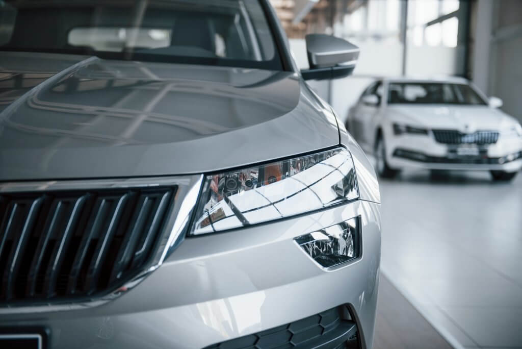 Nahaufnahme der silbernen Fahrzeugfront, dahinter ein weiteres silbernes Auto im hellen Showroom; automatisch gespeicherter Entwurf