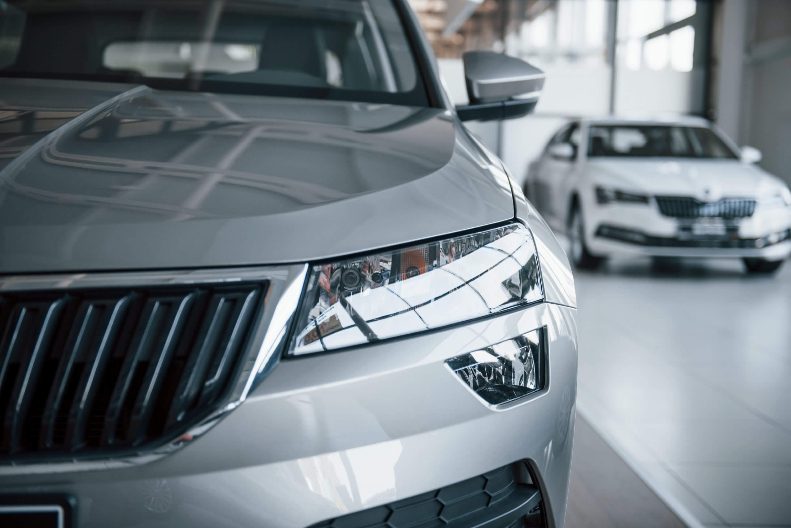 Nahaufnahme der silbernen Fahrzeugfront, dahinter ein weiteres silbernes Auto im hellen Showroom; automatisch gespeicherter Entwurf