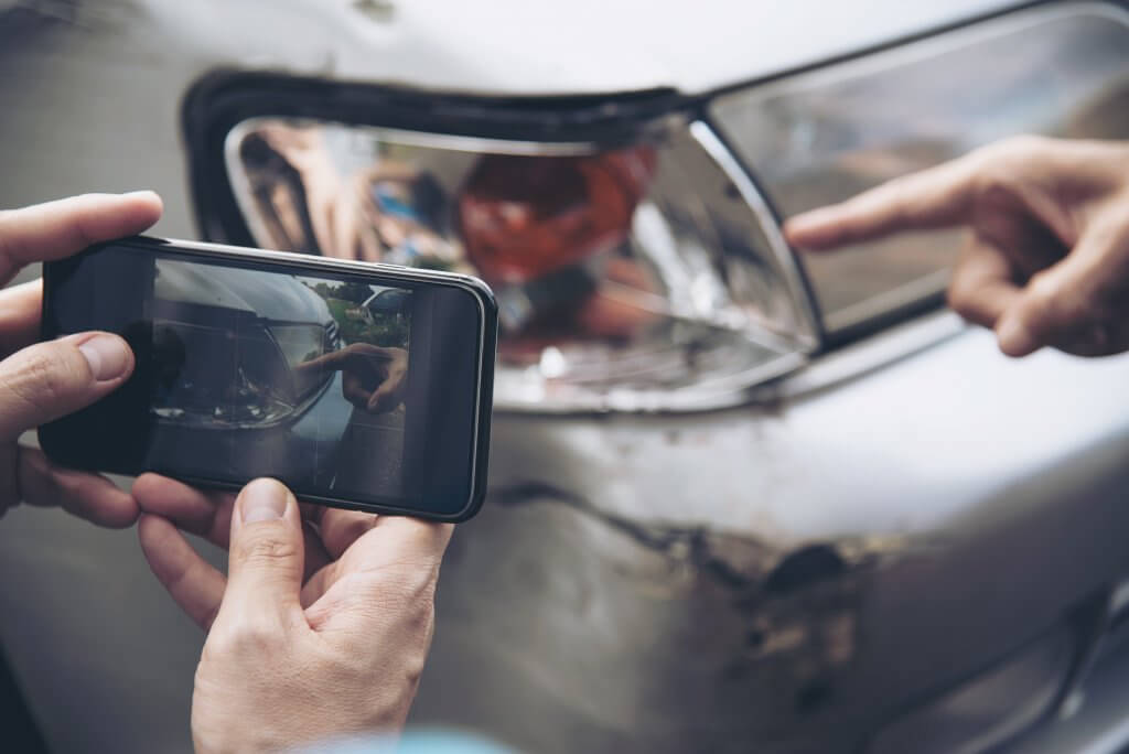 Nahaufnahme von Händen, die mit einem Smartphone eine beschädigte Autotür fotografieren, während jemand auf einen Kratzer zeigt