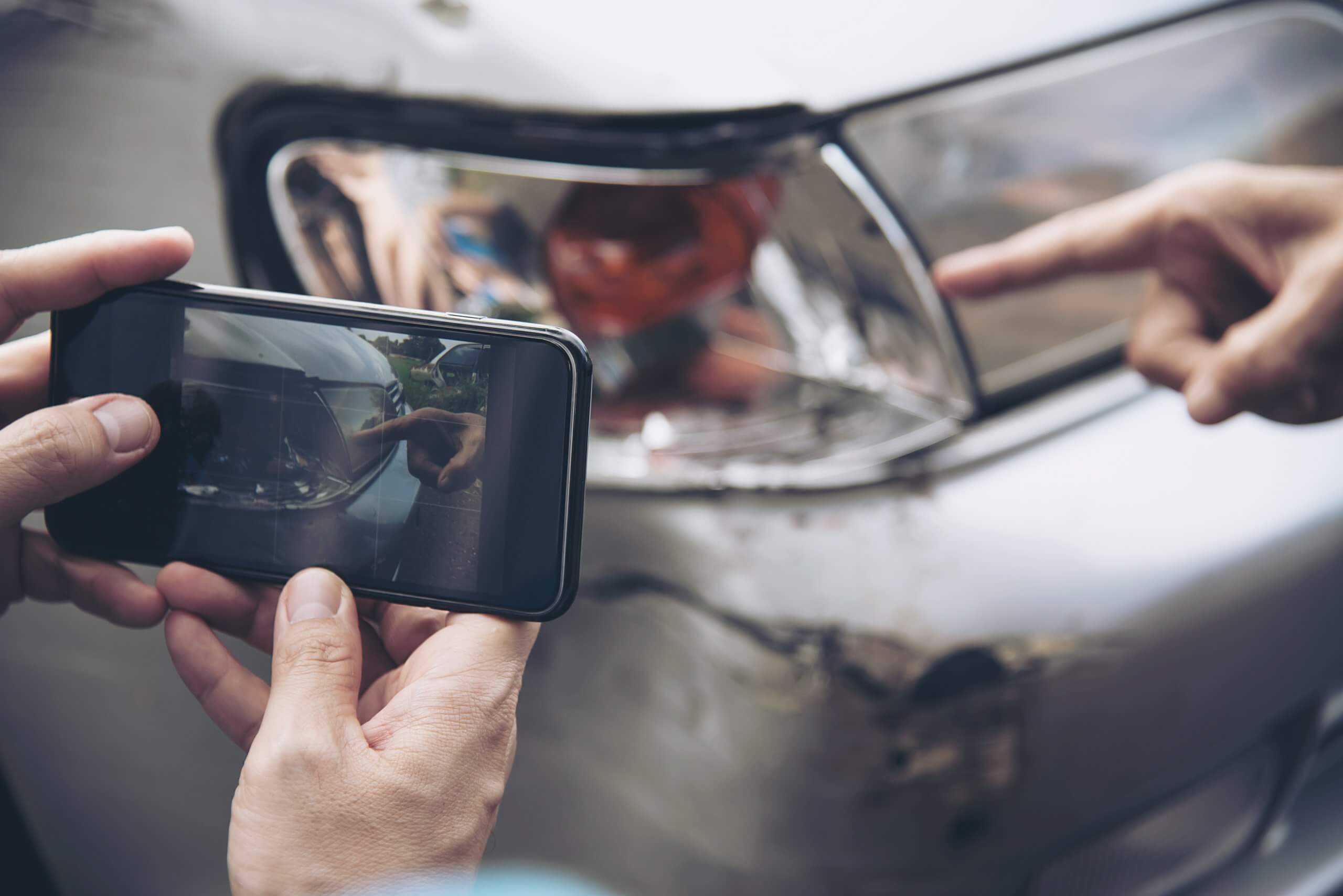 Nahaufnahme von Händen, die mit einem Smartphone eine beschädigte Autotür fotografieren, während jemand auf einen Kratzer zeigt